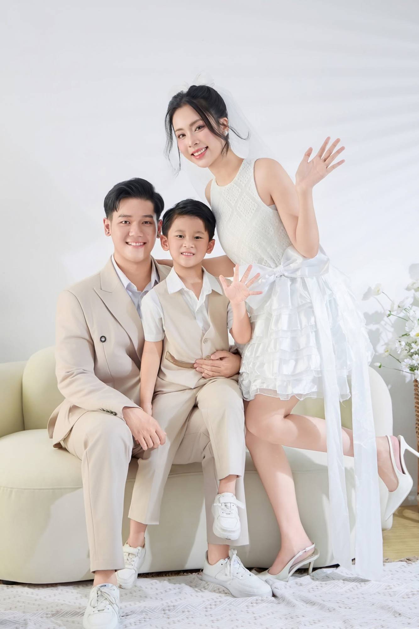 Gia đình ba người mặc tông be ngồi sofa trắng, mẹ và bé vẫy tay